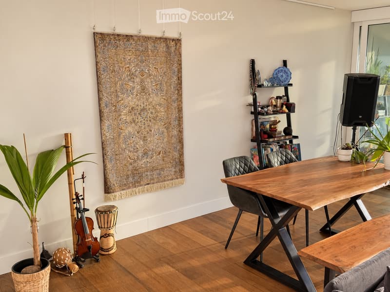 A modern, well-furnished room with a large wooden table, chairs, a shelf with books and decorative items, a speaker, and a large tapestry on the wall. The room also features a potted plant, a drum, and a violin in the corner.