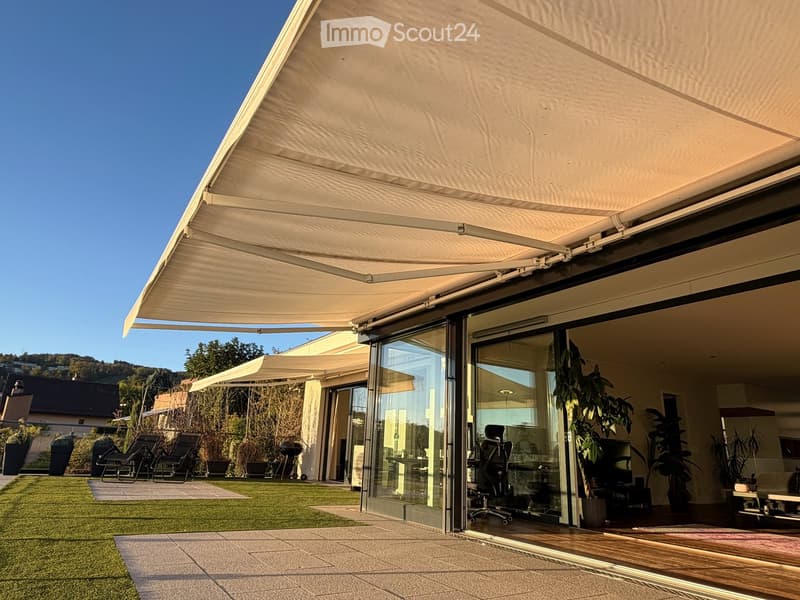 A modern outdoor patio with a large retractable awning, glass sliding doors leading to an indoor space, lounge chairs, a grill, and a well-maintained lawn.