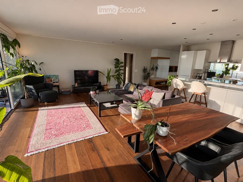 Modern living room with wooden flooring, large windows, grey sofa, coffee table, chairs, and a dining area with a wooden table and chairs.