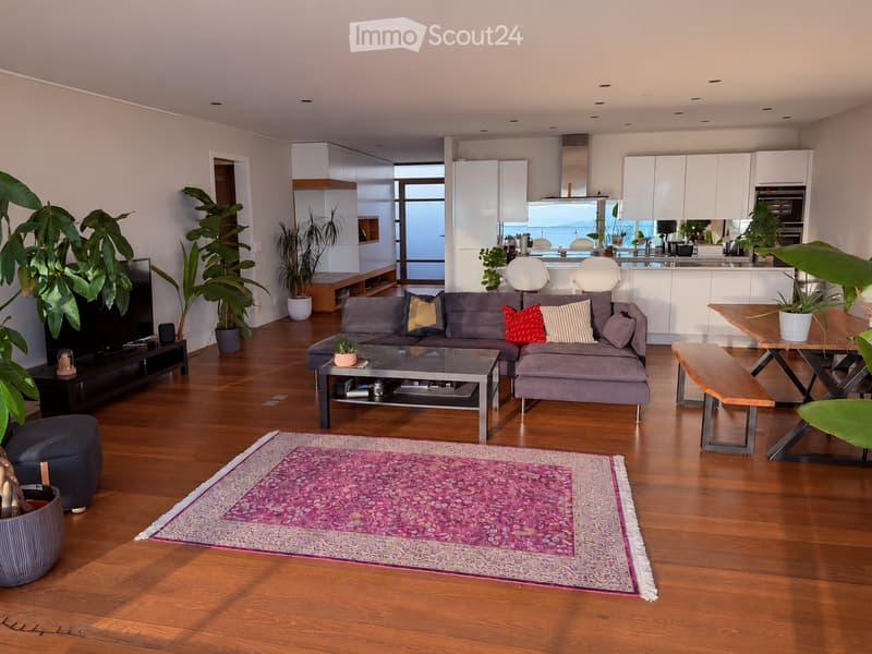 Modern living room with hardwood floors, grey sectional sofa, coffee table, ottoman, dining area with table and bench, large potted plants, and a view of the kitchen area.