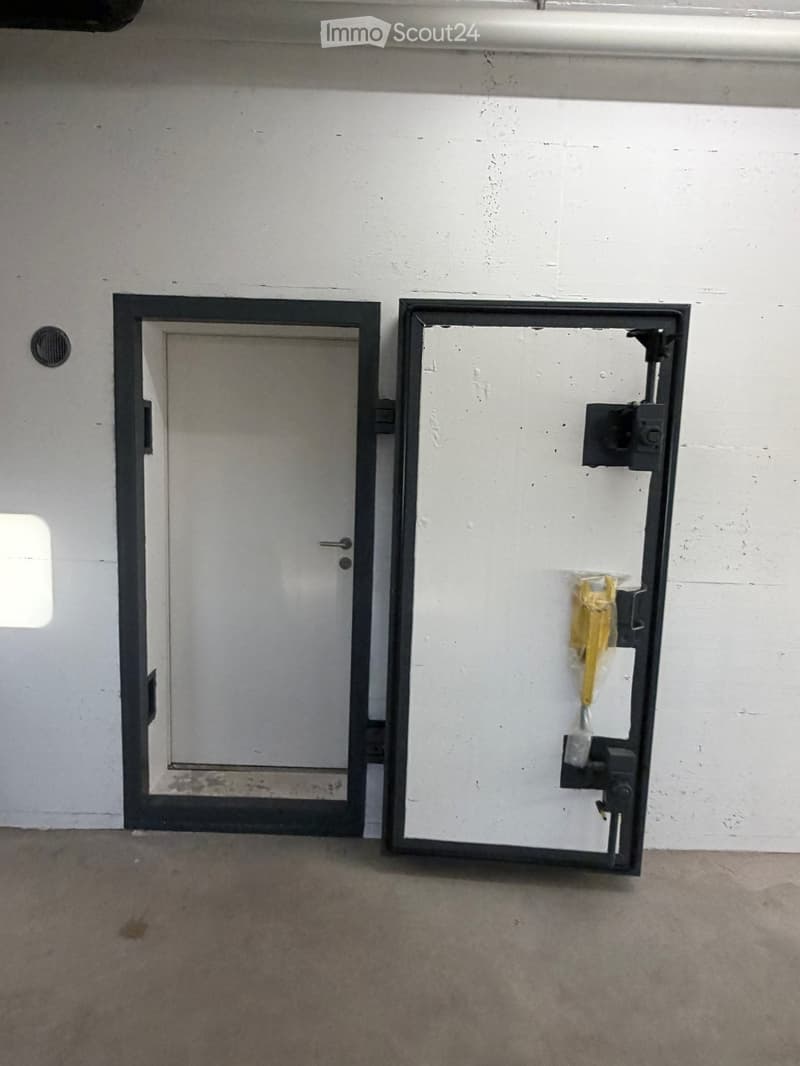 Empty room with a white wall, a closed door, and a partially open window frame with a yellow strap visible.
