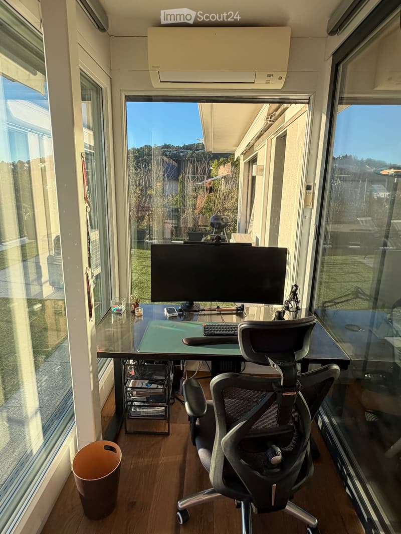 Modern office space with large windows, a desk with a monitor, keyboard, and chair, a trash can, and a storage unit.