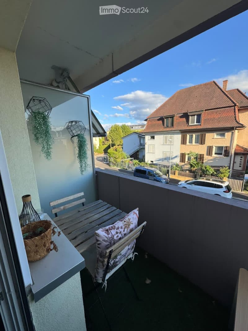 Balcony with a wooden table, a chair with a cushion, a basket on the railing, and hanging plants.