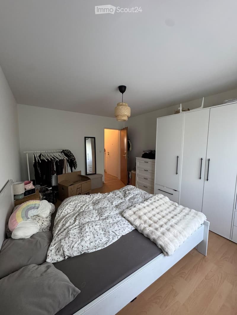 A modern bedroom with a neatly made bed, a large wardrobe, a mirror, and a clothing rack. The room features a hanging lamp and a small nightstand with a lamp.