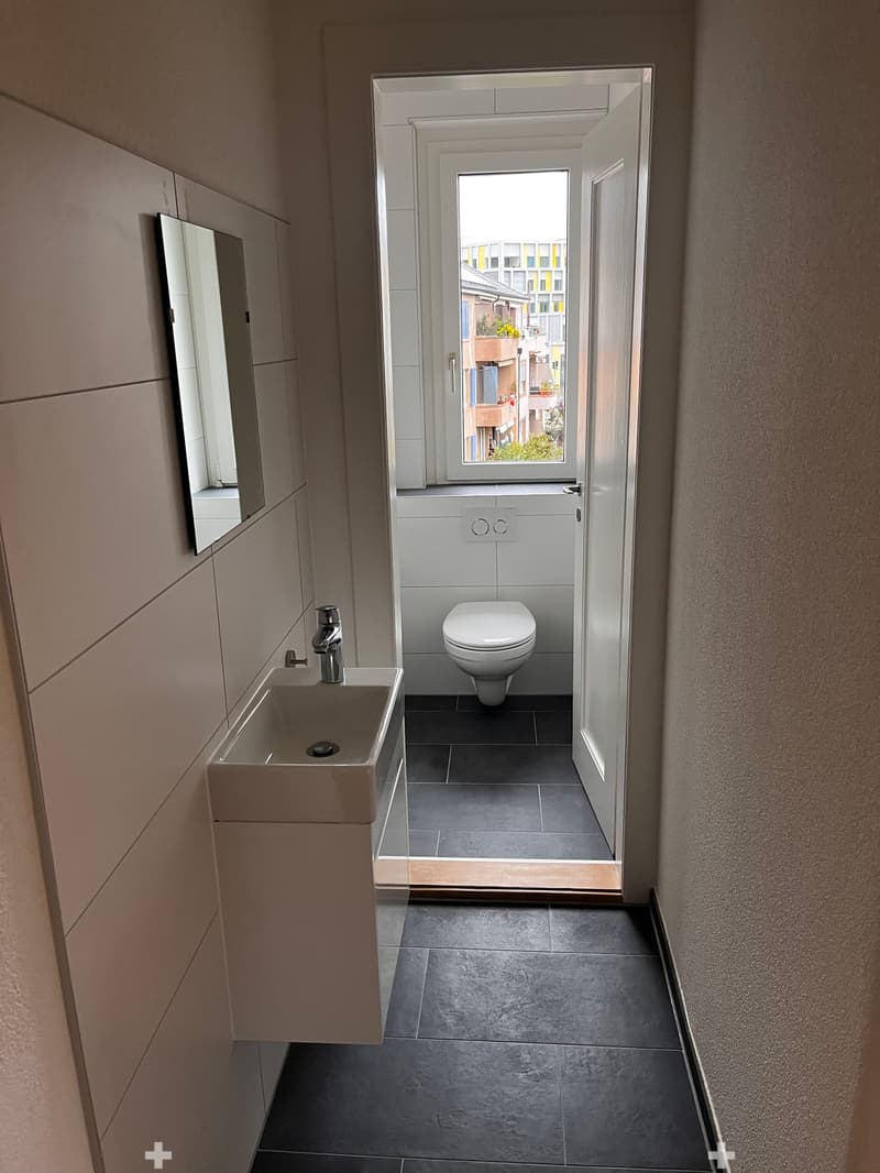 Modern bathroom with a sink, faucet, toilet, and mirror. The space features dark tiled flooring and light-colored walls.