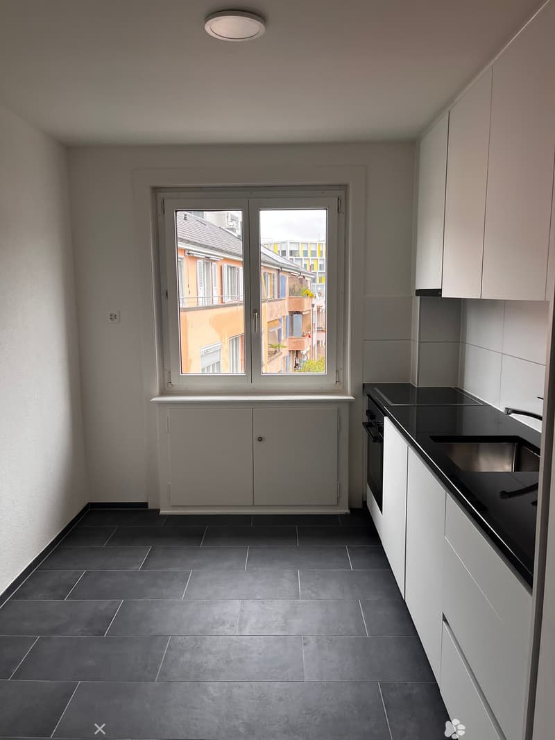 Modern kitchen with white cabinets, black countertop, and dark tiled floor. Features a large window with a view of the street.
