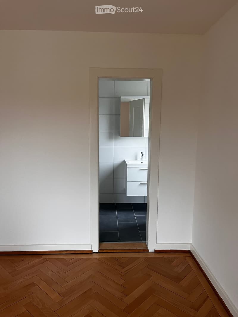 Empty room with wooden herringbone flooring and white walls, featuring an open doorway leading to a modern bathroom with white tiles, a sink, and a mirror.