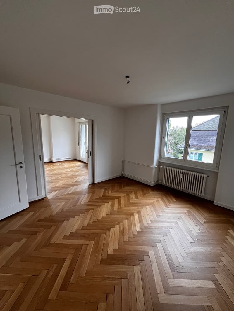 Empty room with wooden herringbone flooring, white walls, and a radiator. There are two doors and a window with a view of a neighboring building.