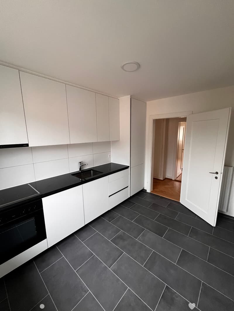 Modern kitchen with white cabinets, black countertops, and dark tiled flooring. Features a sink, faucet, and built-in oven.