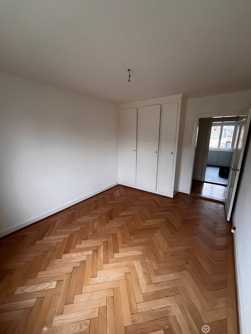 Empty room with herringbone wooden flooring, white walls, and a set of white built-in wardrobes. A doorway leads to another room with a visible tiled floor.