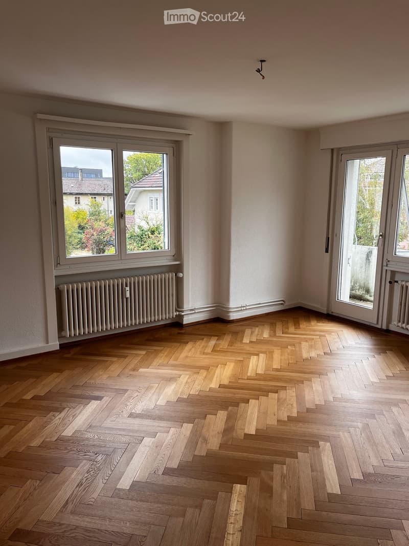 Empty room with wooden herringbone flooring, large windows, radiators, and white walls.