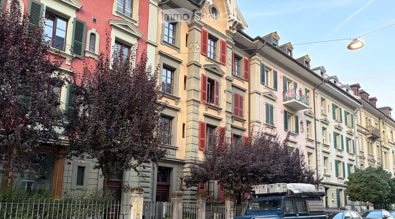 A row of multi-story residential buildings with colorful facades, green shutters, and ornate architectural details, featuring balconies and trees lining the street.