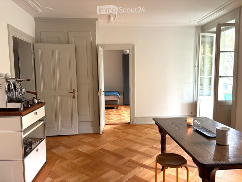 Bright, open-plan room with wooden herringbone flooring, a wooden dining table with a stool, a coffee machine on a counter, and an open doorway leading to a bedroom area with a bed.