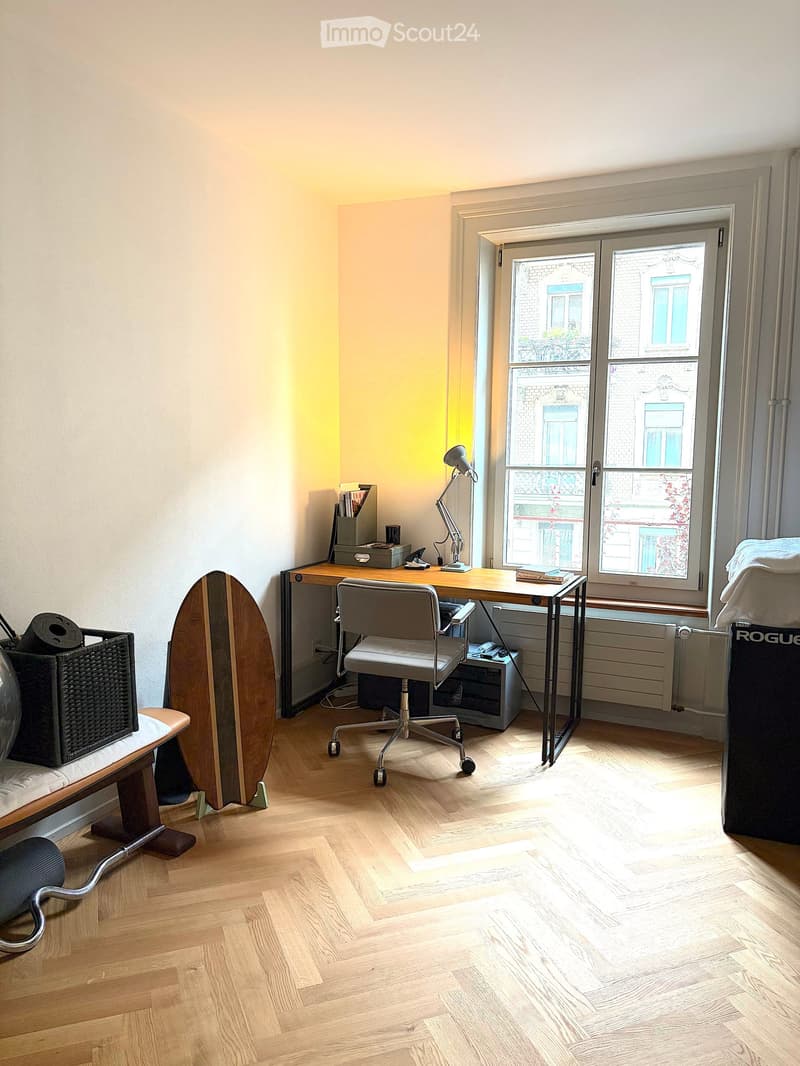 A well-lit room with wooden flooring, a desk with a chair, a lamp, a surfboard leaning against the wall, a small table with a basket, and a window with a view of the street.