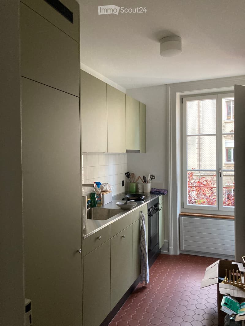 The kitchen features light-colored cabinets, a stainless steel sink, an oven, and a window with a view of a brick building. The floor is tiled in a hexagonal pattern, and various kitchen utensils and items are visible on the counter.