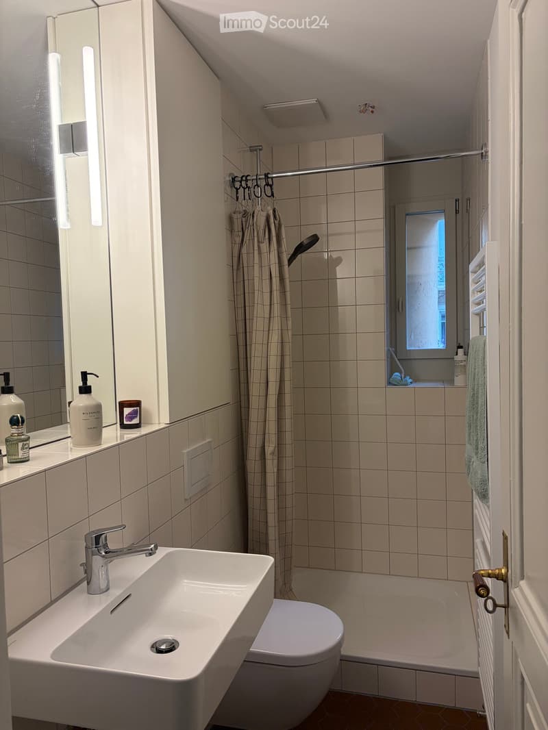 Modern bathroom with a white sink, chrome faucet, toilet, tiled walls, and a shower area with a curtain. A mirror with toiletries is mounted on the wall, and a towel hangs on a rack.