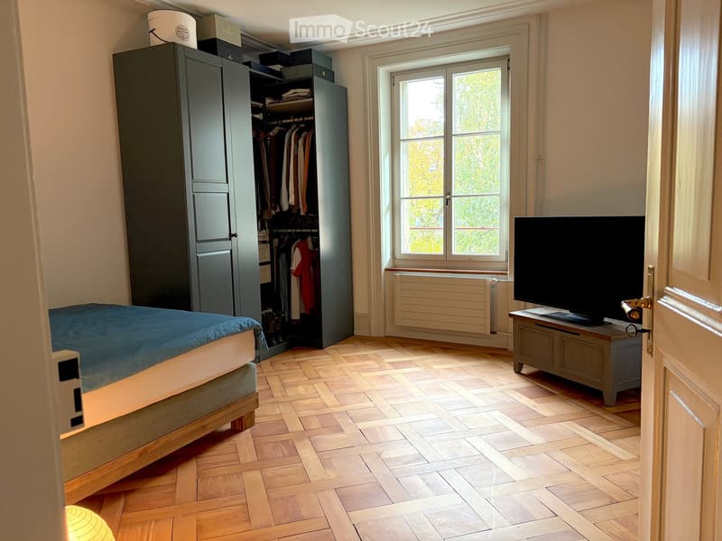 Bright bedroom with wooden herringbone flooring, large window, bed with blue bedding, wardrobe with hanging clothes, TV on a stand, and a small table.