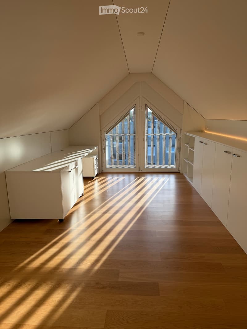 Bright, empty attic room with wooden flooring, white built-in cabinets, and large double doors leading to a balcony.