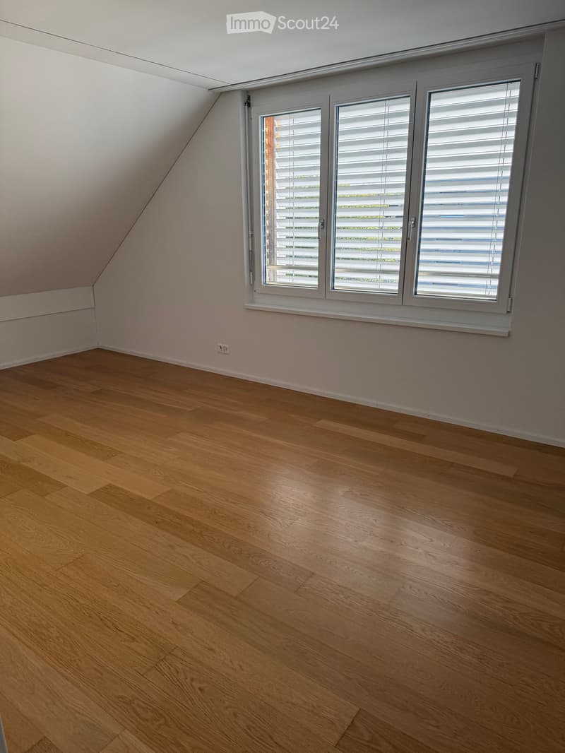 Empty attic room with wooden flooring, white walls, and a large window with blinds.