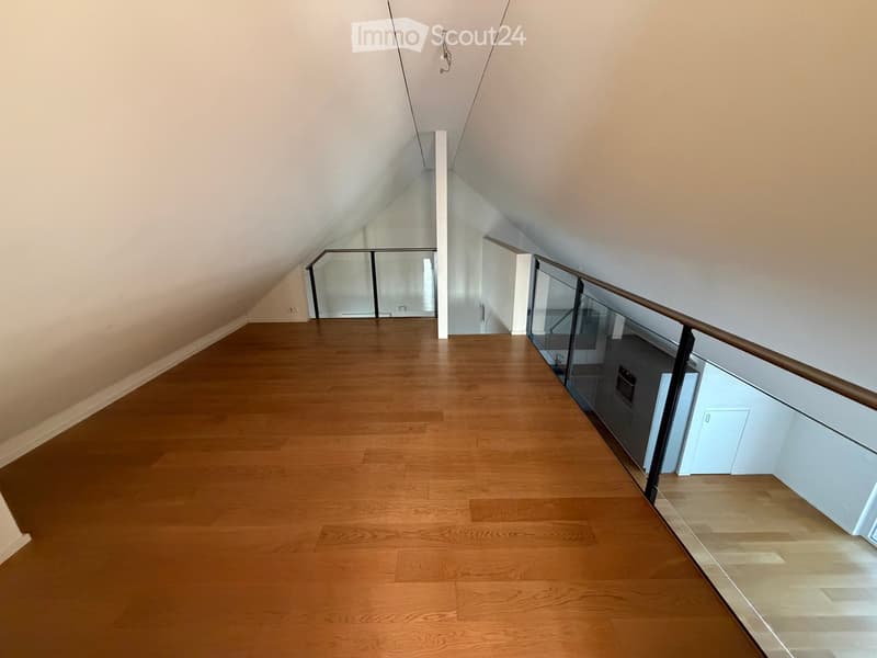 Spacious, well-lit hallway with wooden flooring and glass railings leading to an open area. The walls are painted in a light neutral color, and the ceiling has a sloped design typical of an attic space.