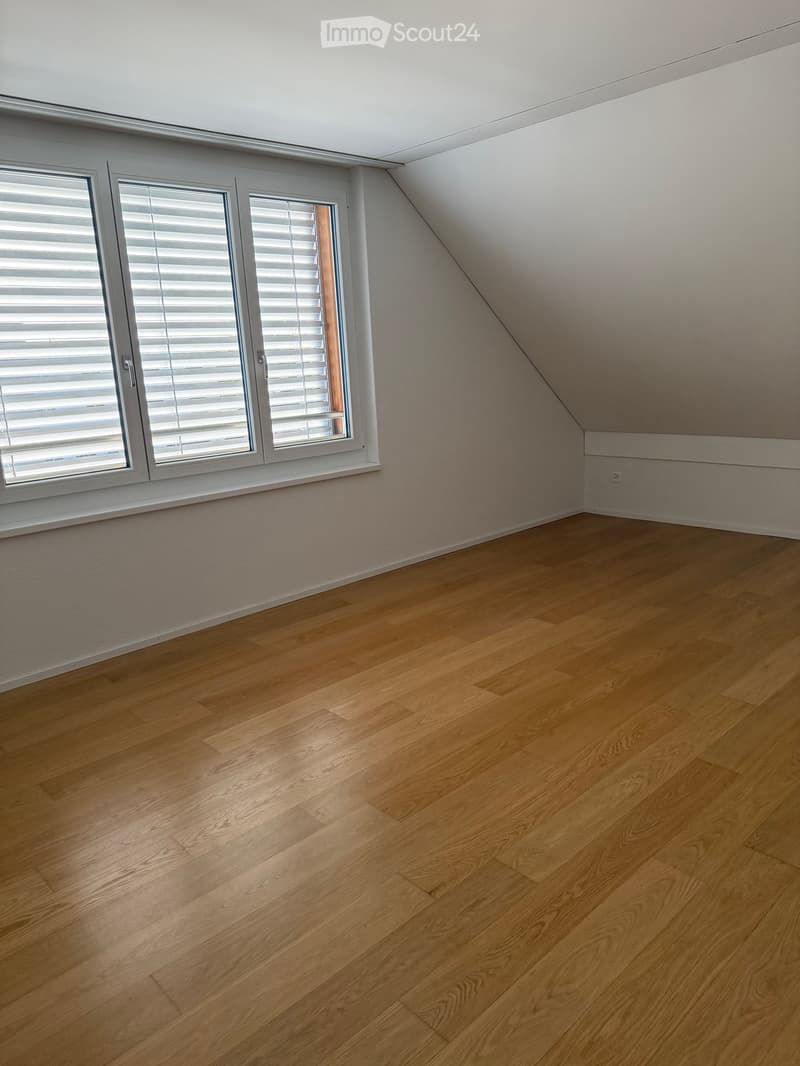Empty room with wooden flooring, white walls, and large windows with blinds. The ceiling has a sloped design typical of an attic space.
