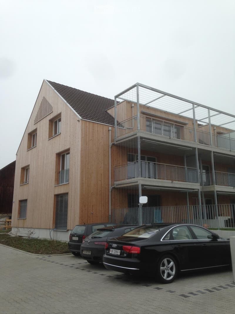 Modern two-story building with wooden facade and metal balcony, with three parked cars in front.