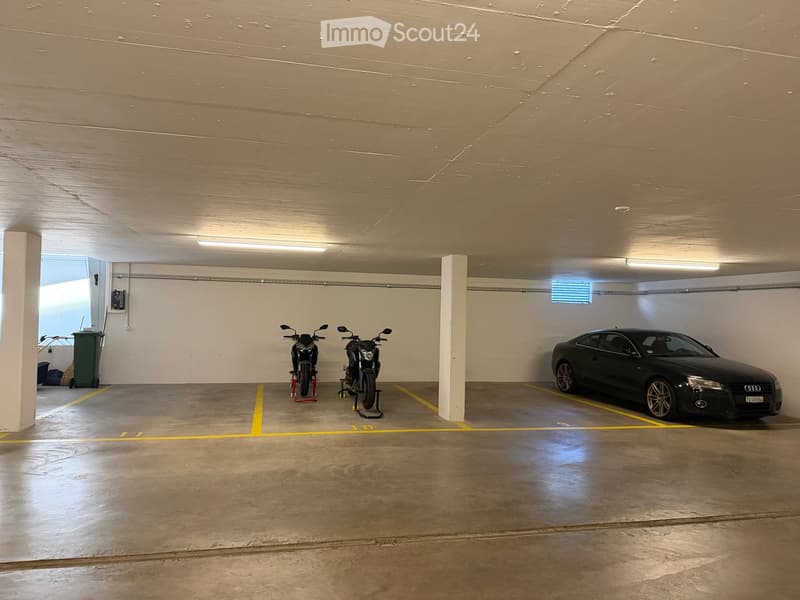 Underground parking area with a car and two motorcycles parked in designated spots, concrete floor, white walls, and fluorescent lighting.