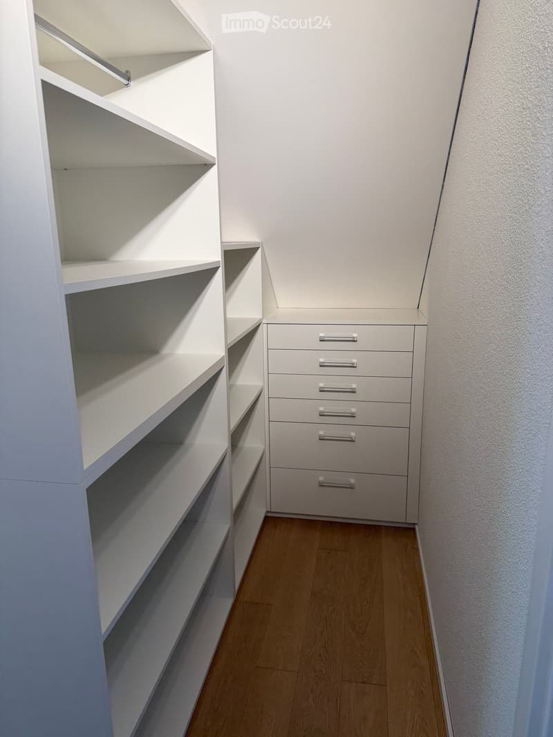 Empty closet with white built-in shelves and drawers, light wooden floor, white walls.