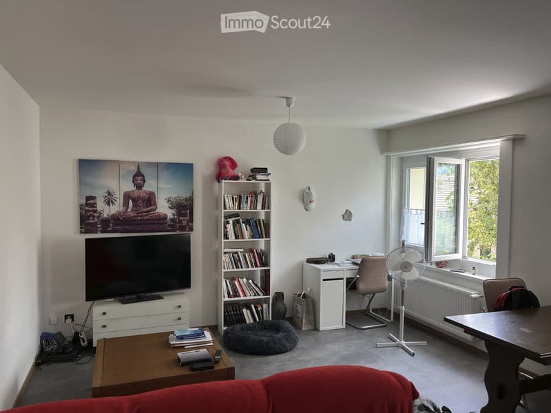 A modern living room with a large TV, bookshelf, coffee table, sofa, and a fan near the window.