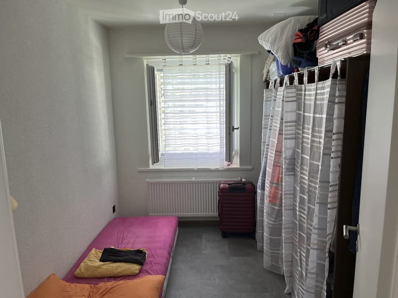 Small bedroom with a single bed, pink and orange bedding, a suitcase, a radiator, and a curtained closet.