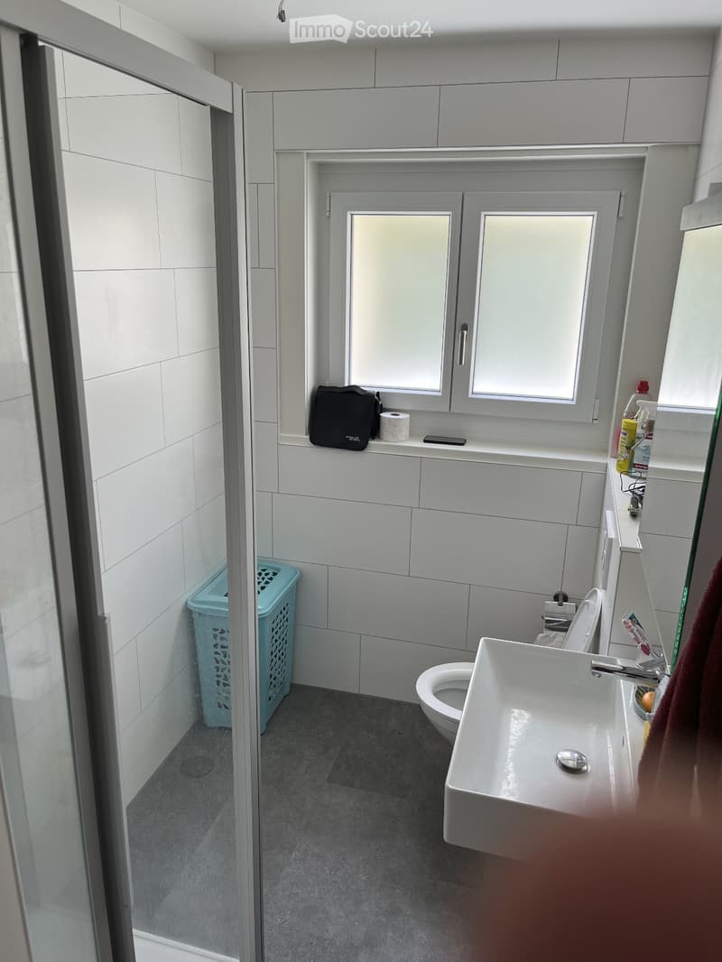 Modern bathroom with white tiles, a sink, toilet, and a window above the sink. A towel hangs on the side, and a laundry basket is placed near the shower area.