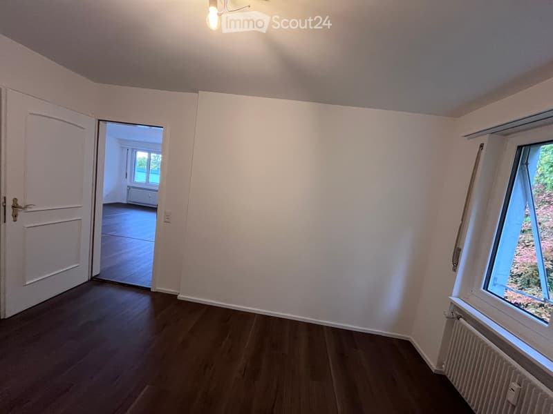 Empty room with wooden flooring, white walls, and a large window with a view of greenery. A door leads to another room with similar flooring and white walls.