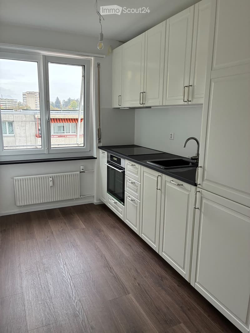 Modern kitchen with white cabinets, black countertop, sink, faucet, and oven. Wooden flooring and large windows provide natural light.