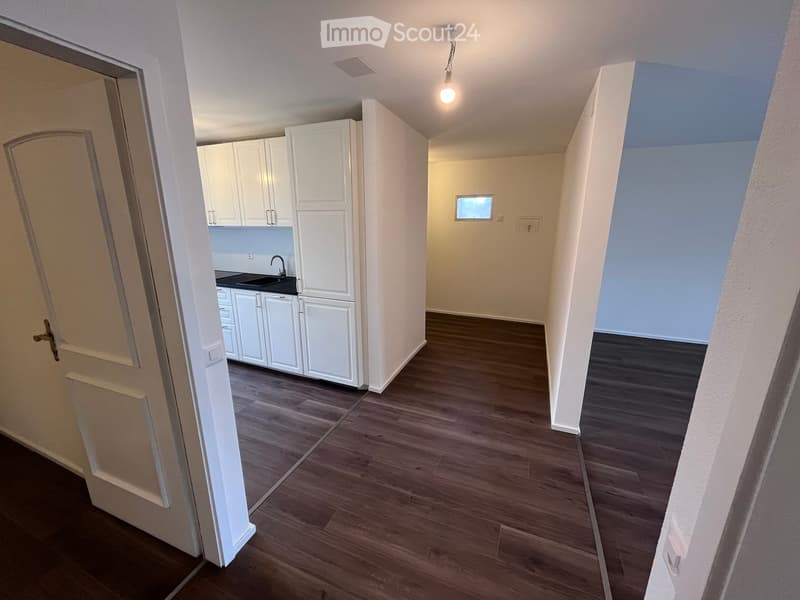 Empty hallway with wooden flooring, white walls, and a small window. A kitchen area is visible through an open doorway, featuring white cabinets and a sink.