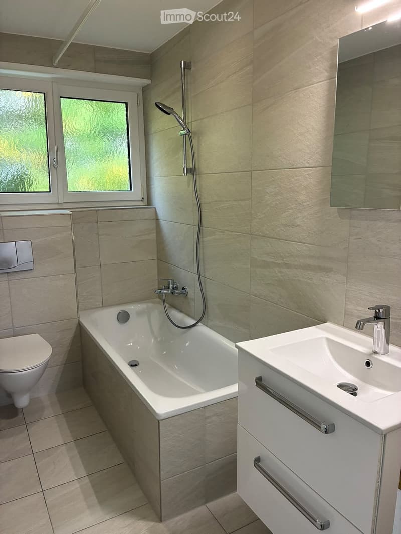 Modern bathroom with a bathtub, toilet, sink, and large mirror. Features a window with frosted glass and a sleek, contemporary design.