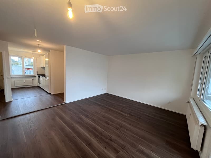 Empty room with wooden flooring, white walls, and large windows. The adjacent kitchen area is also visible with white cabinets and a countertop.