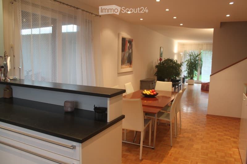 Modern kitchen and dining area with a black bar counter, white chairs, wooden dining table, and a bowl of fruit. The space features light wooden flooring, white walls, and large windows with sheer curtains.