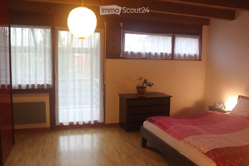 Cozy bedroom with wooden flooring, large windows with white curtains, a bed with red and white bedding, a nightstand with a lamp, a drawer unit, and a small potted plant on top.