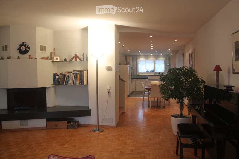 Open-plan living and dining area with wooden flooring, fireplace, bookshelf, floor lamp, piano, dining table, chairs, and potted plant.