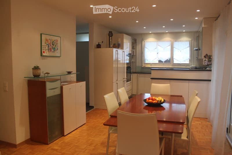 Modern kitchen and dining area with wooden flooring, white cabinets, a dining table with chairs, and a bowl of fruit on the table.