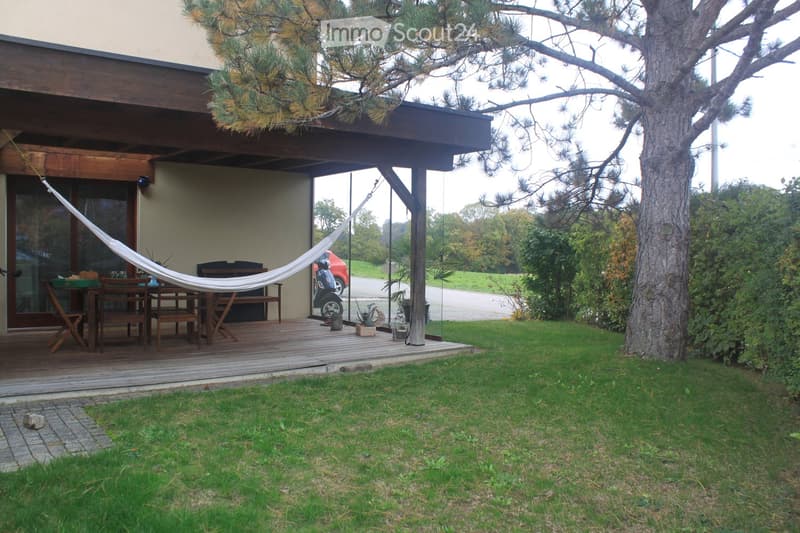 A wooden deck area with a hammock, a small table and chairs, and a view of a grassy lawn and surrounding trees.