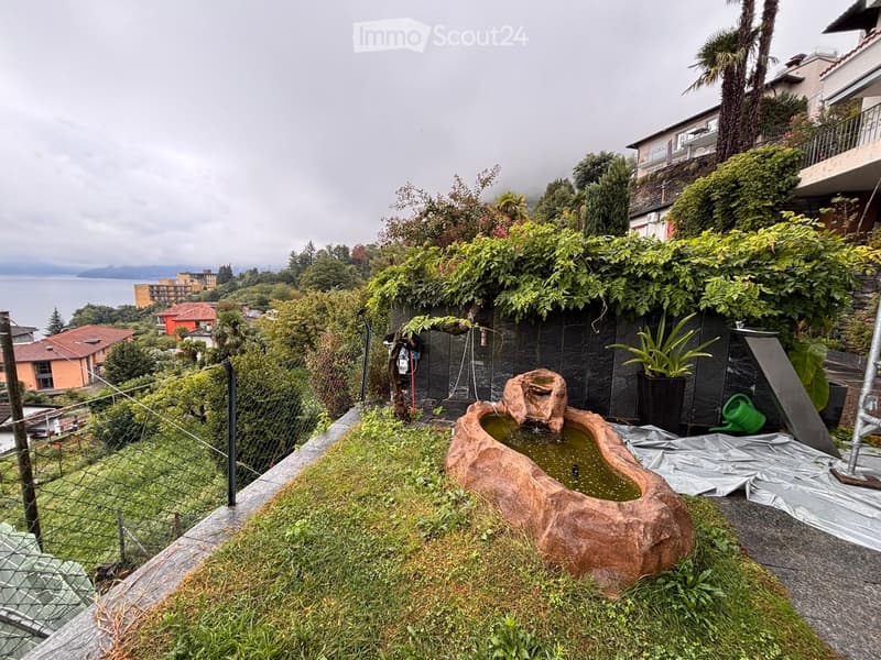A lush garden area with a stone water feature, surrounded by greenery and a view of the lake and distant buildings.