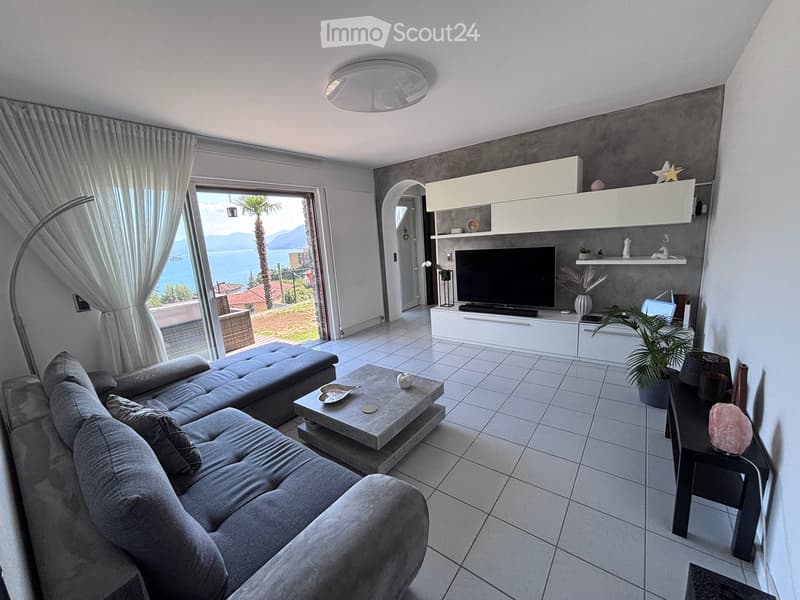 Modern living room with a grey sectional sofa, coffee table, TV, floor lamp, and a view of the sea through large windows.