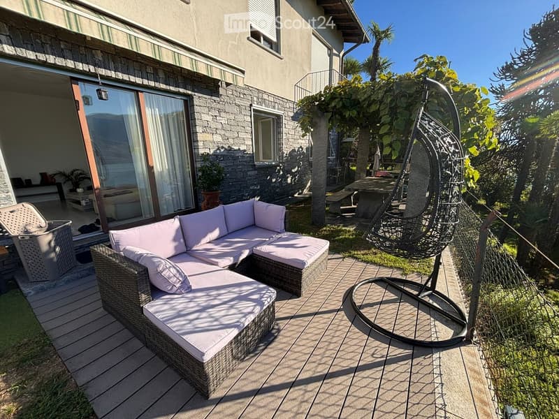 Outdoor patio with a wicker sofa, hanging egg chair, and a table surrounded by lush greenery and a modern house in the background.