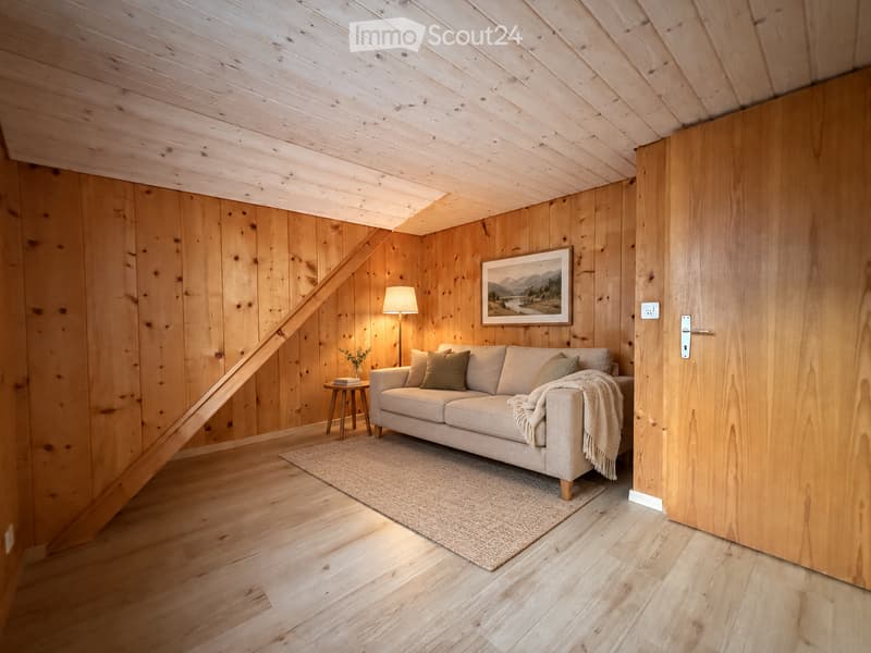 Cozy living room with wooden walls and ceiling, featuring a light-colored sofa, throw blanket, small wooden stool, table lamp, and a framed picture on the wall.