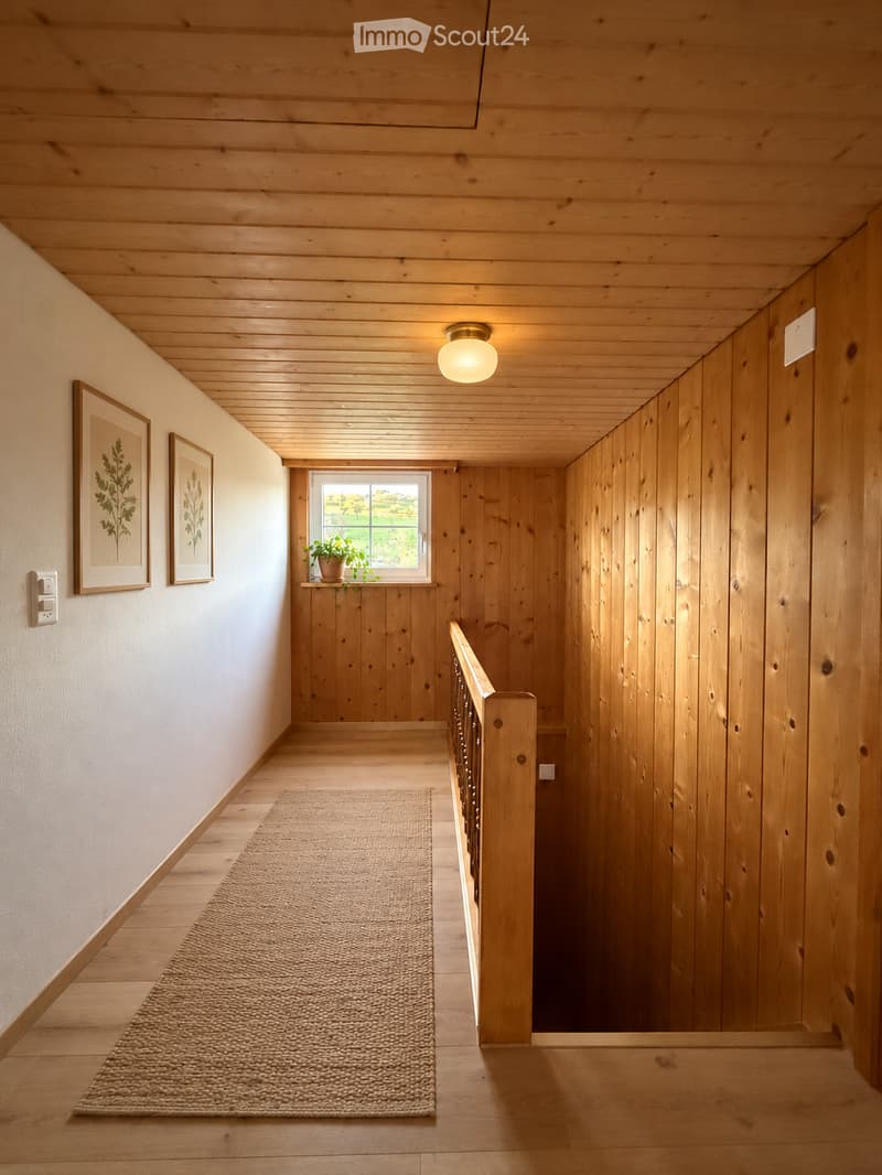Wooden hallway with a staircase, light fixture, framed artwork, small potted plant on a windowsill, and a textured rug on the floor.