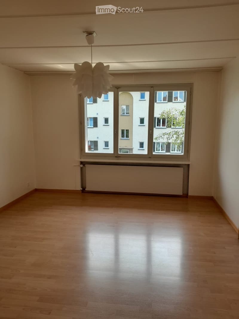 Empty room with wooden flooring, large windows, radiator, and a hanging light fixture.