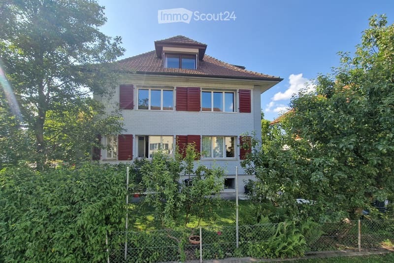 Two-story house with white brick facade, red shutters, and multiple windows, surrounded by lush greenery and a fence.