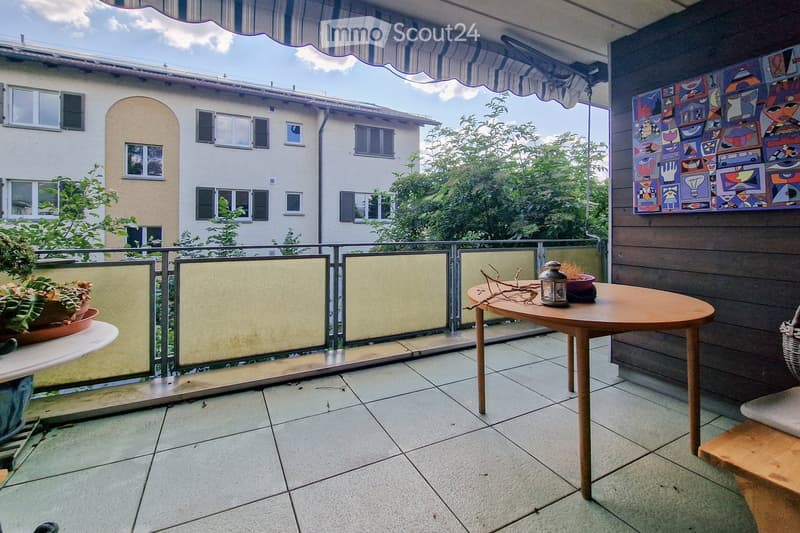 Balcony with tiled floor, round wooden table, chair, lantern, and potted plants. Features a striped awning and a colorful abstract painting on the wall.