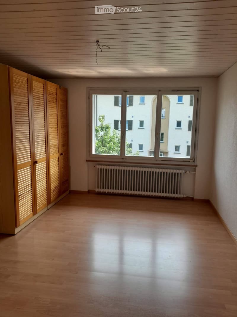 Empty room with wooden flooring, large window with view of buildings, radiator, and wooden closet with louvered doors.